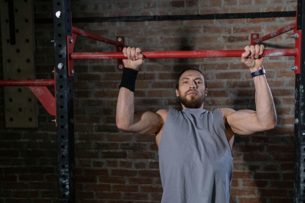 Muscular man doing pull-up exercise indoors on a red bar against a brick wall.