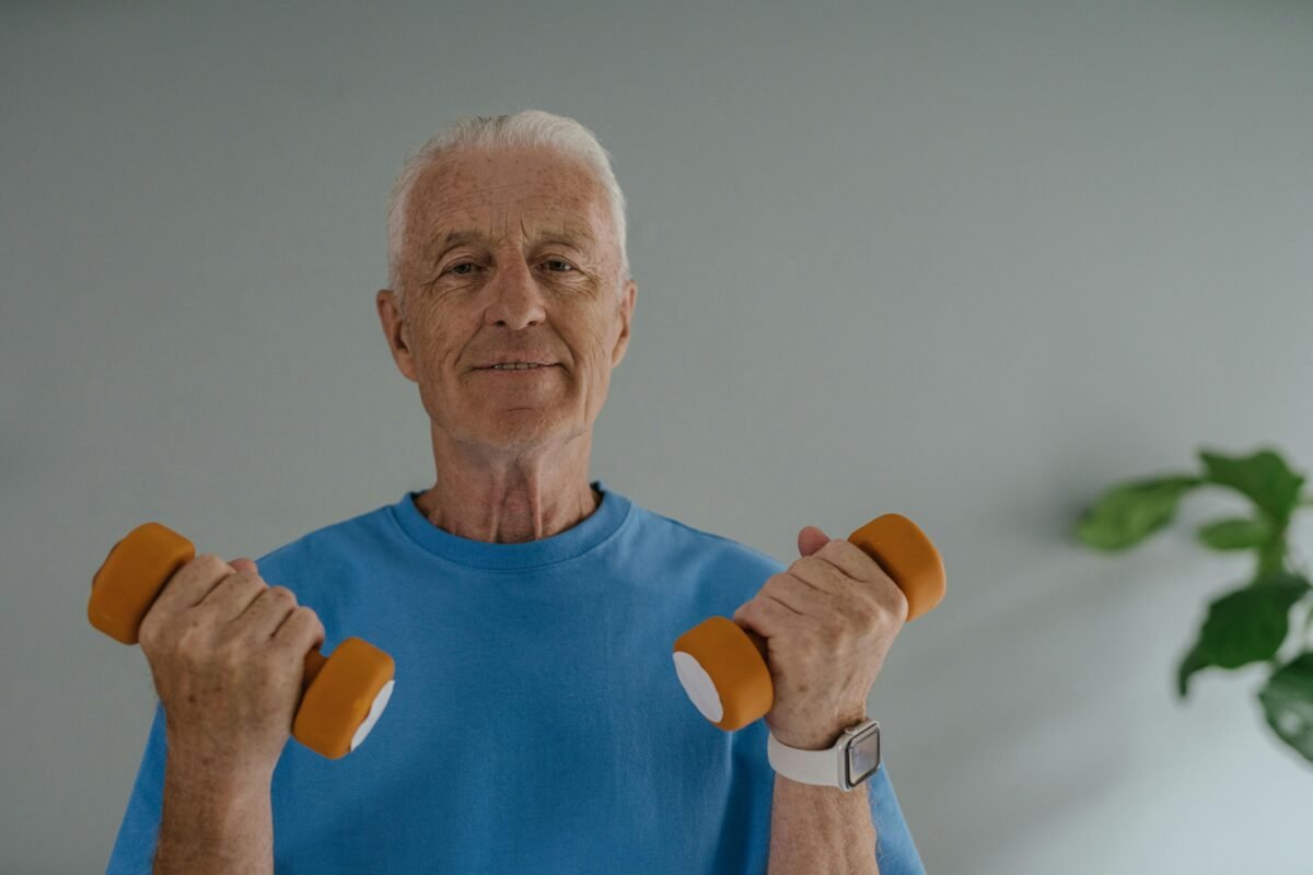 Elderly man in blue shirt lifting dumbbells and smiling indoors, promoting active lifestyle.